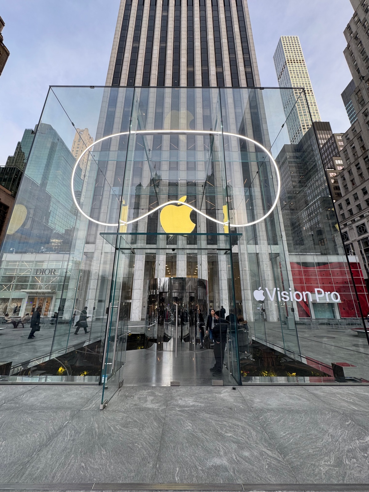 The glass cube of the Apple Store on Fifth Avenue at dusk with the Apple logo inside it and a white Vision Pro outline drawn around the logo
