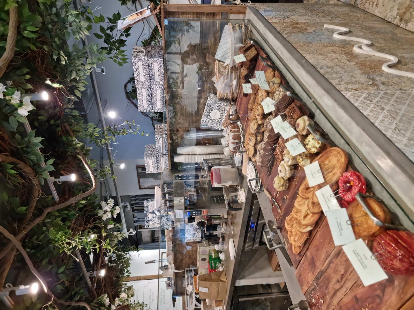 A bakery counter with pastries laid out on a stone slab beneath a dense canopy of artificial greenery and hanging light bulbs