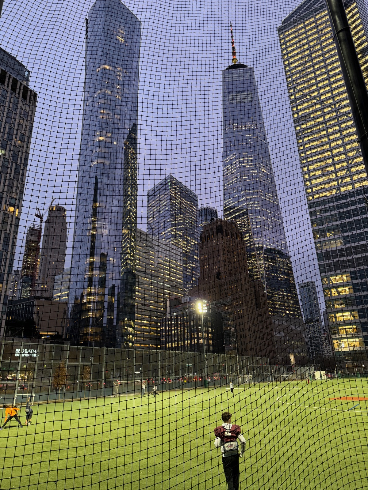 A floodlit green ball field seen through black netting, a player in catcher's gear standing with his back to the camera and One World Trade Center and other towers rising directly behind
