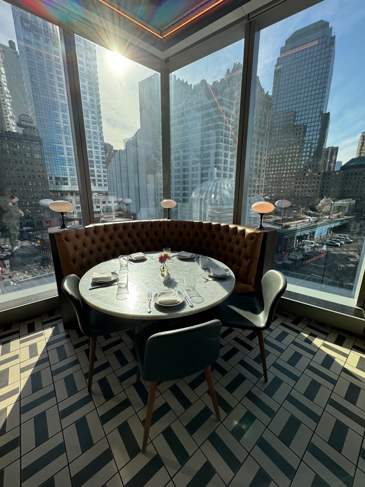 A round table in a curved green banquette in a corner window, patterned tile floor, with glass towers filling both windows