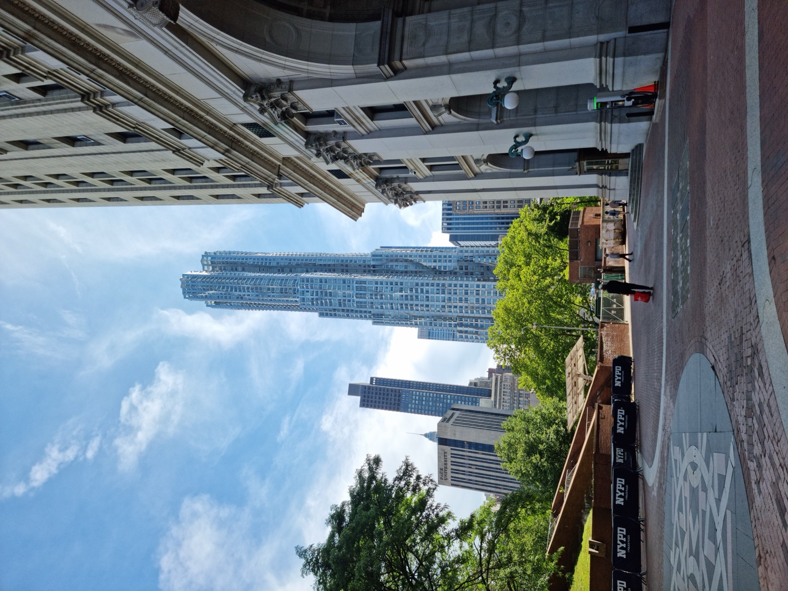 A paved plaza at the southern tip of Manhattan in full summer, heavy green trees on the left and a tall residential tower and stone building beyond