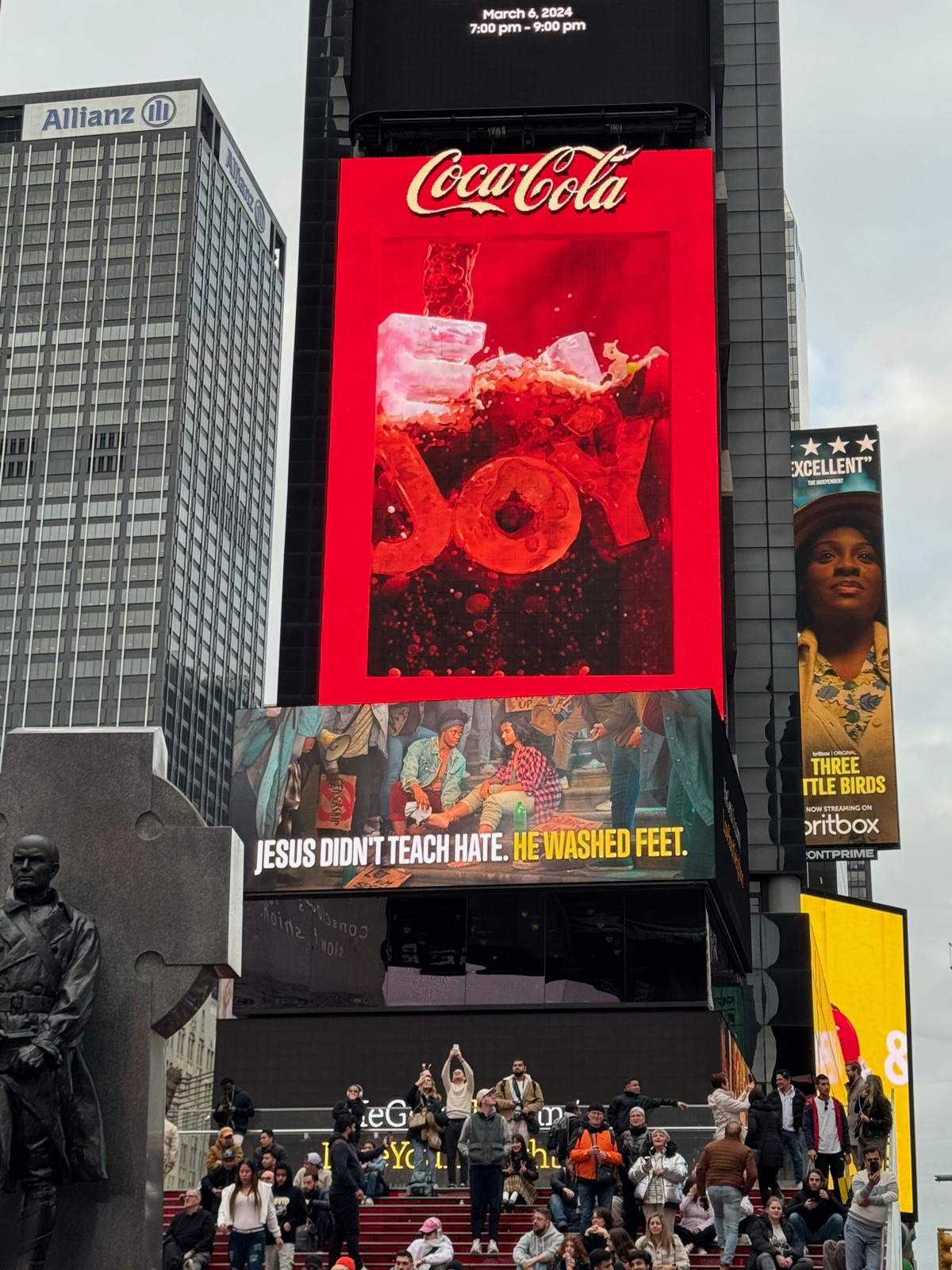 A painted billboard in Times Square showing people on a stoop washing each other's feet, captioned JESUS DIDN'T TEACH HATE. HE WASHED FEET., beneath a large red Coca-Cola screen