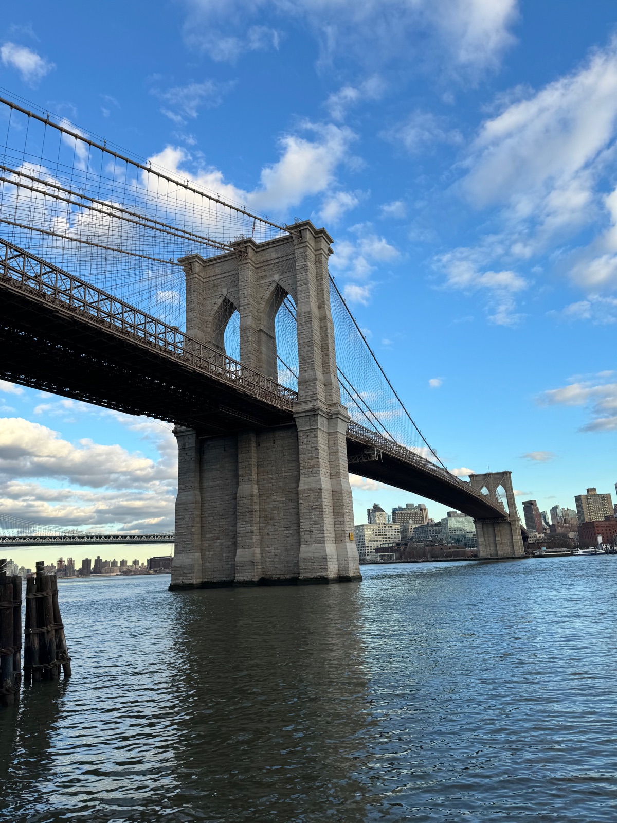 A stone tower of the Brooklyn Bridge rising from the East River under a blue sky with scattered white clouds