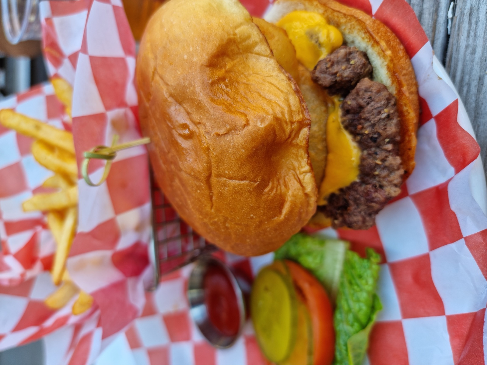 A cheeseburger and fries in a red and white checked paper liner in a basket, with ketchup and pickles on the side