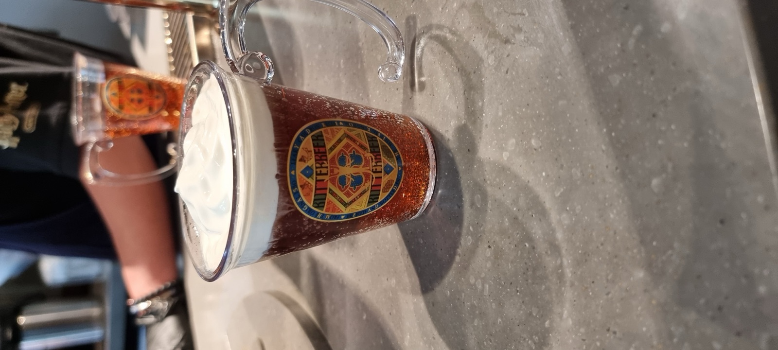A tall glass of amber butterbeer topped with thick white foam on a stone counter, a second glass behind it