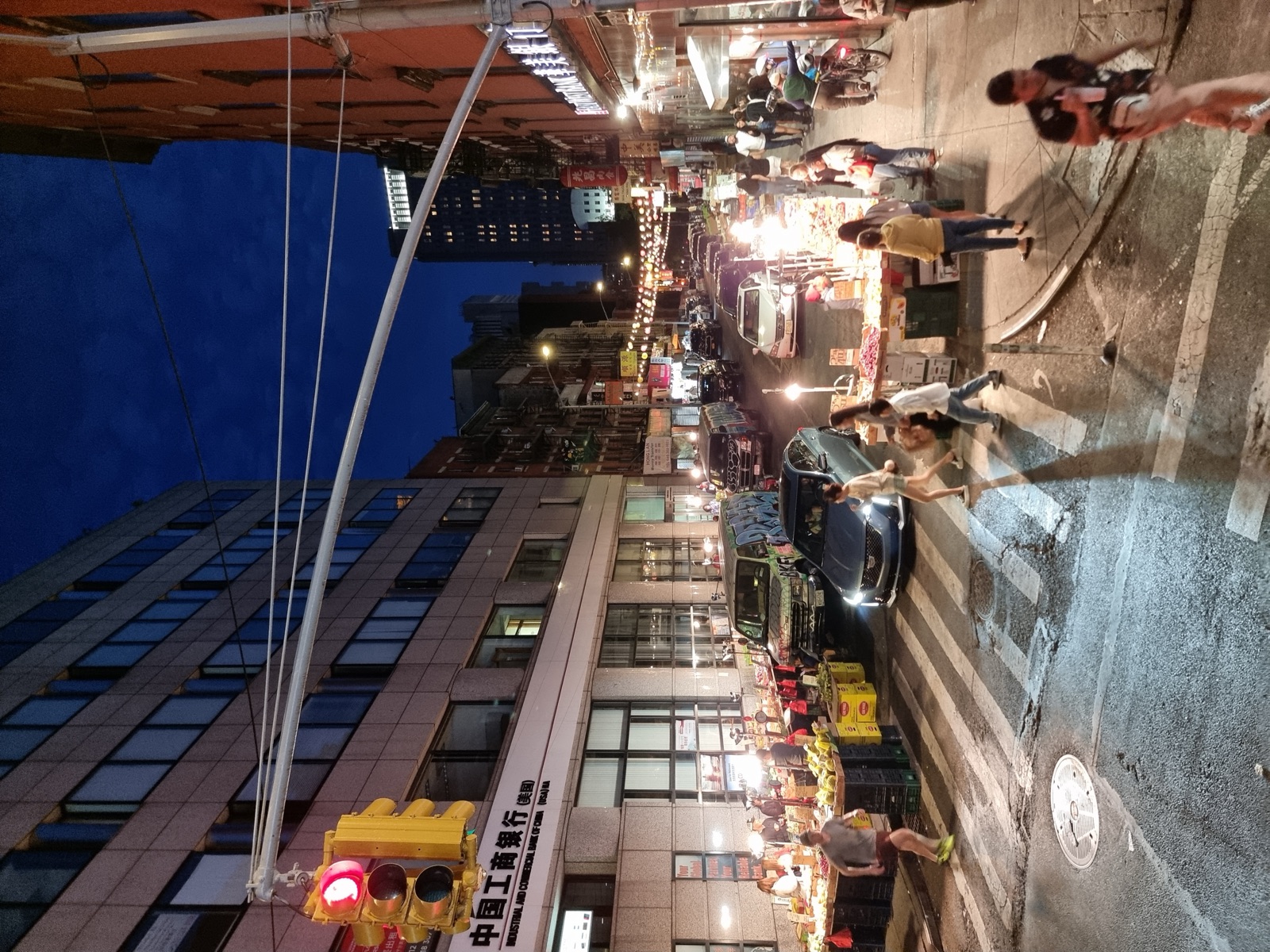 A Chinatown street at night with market stalls of fruit under awnings, strung lights overhead, cars at a crossing and Chinese lettering on the storefronts