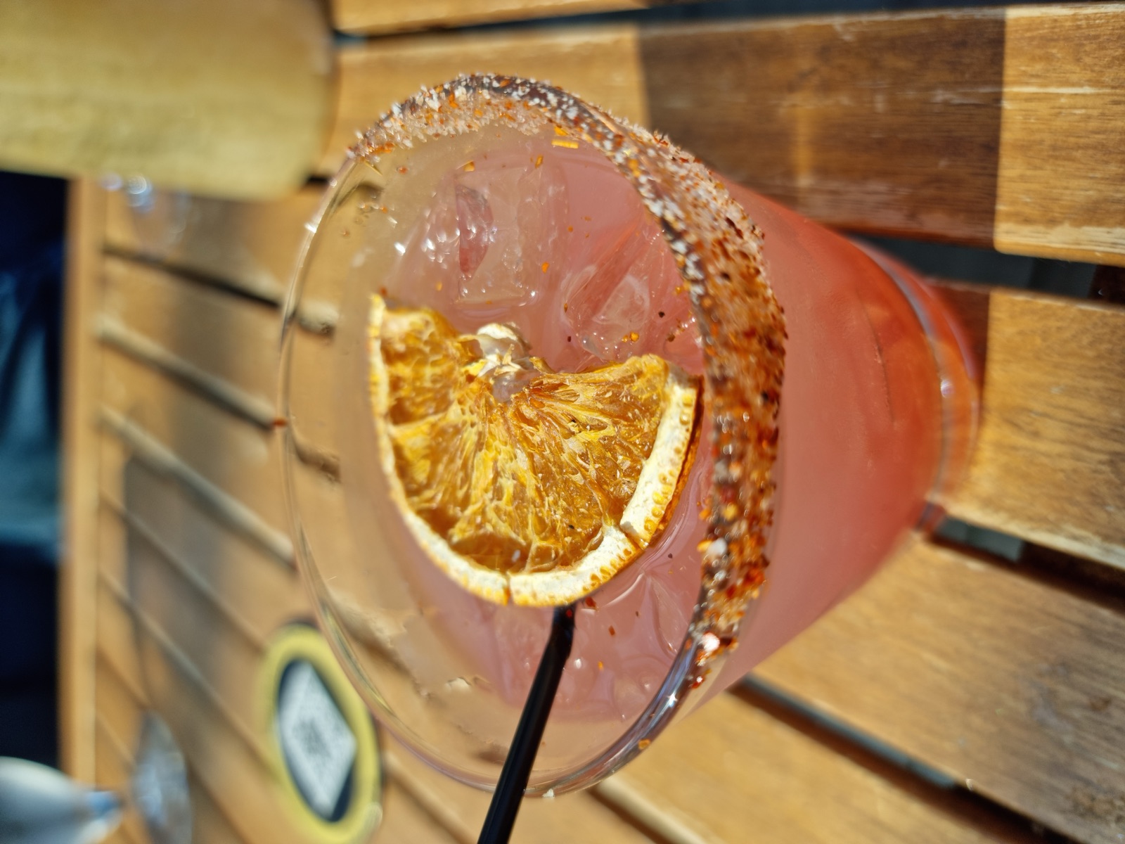 A pink cocktail in a tumbler with a spiced rim and a dried orange wheel, on a slatted wooden table outdoors