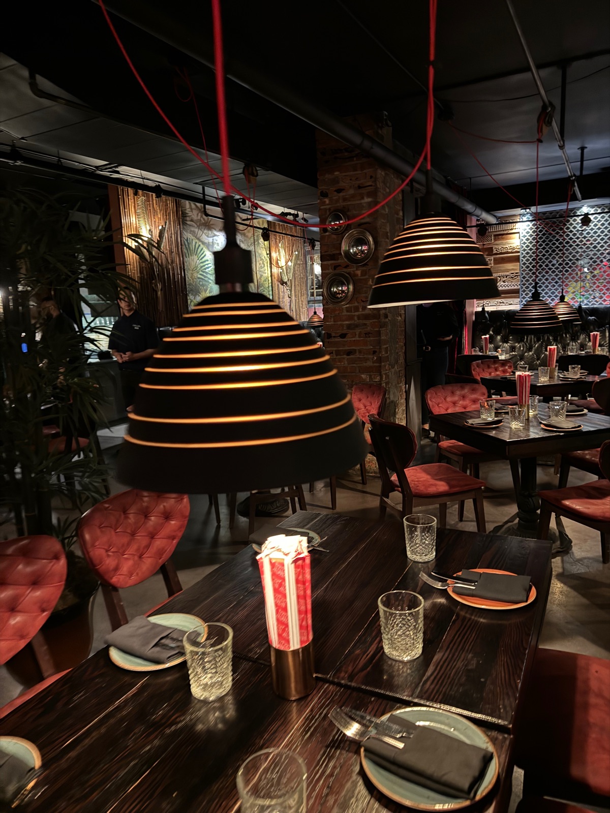 A dark restaurant interior with ribbed copper pendant lamps hanging low over a wooden table, red buttoned banquettes and a brick wall