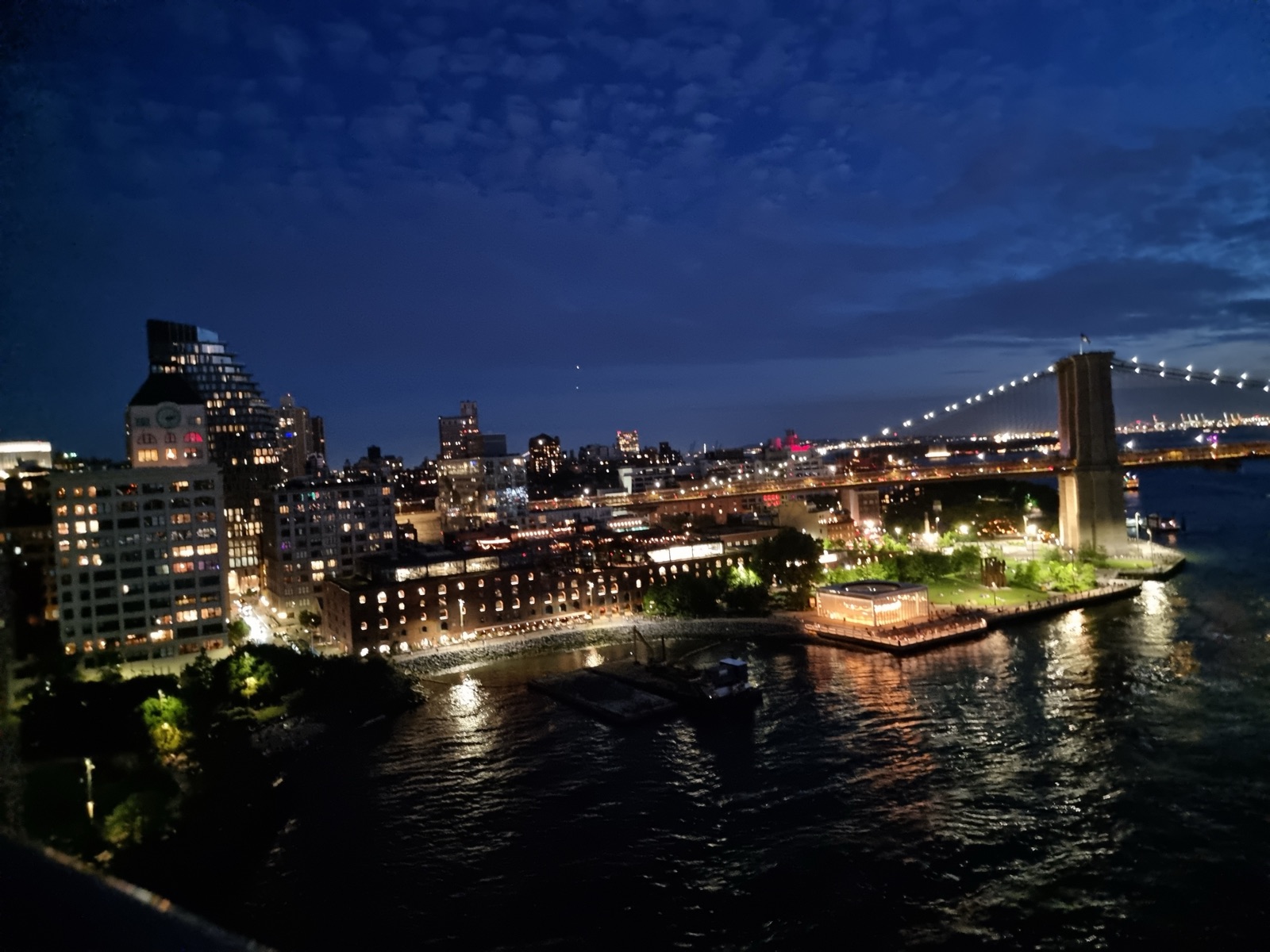 DUMBO in Brooklyn seen from high on the Manhattan Bridge at blue hour, the lit clock tower and brick Empire Stores warehouses along the waterfront, a glowing glass pavilion on the pier, and the Brooklyn Bridge tower with lit cables at the right