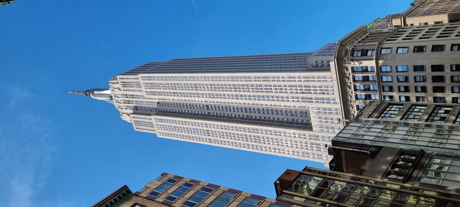 The Empire State Building seen from the street below, its limestone setbacks and mast rising into a clear blue sky above the lower buildings around it