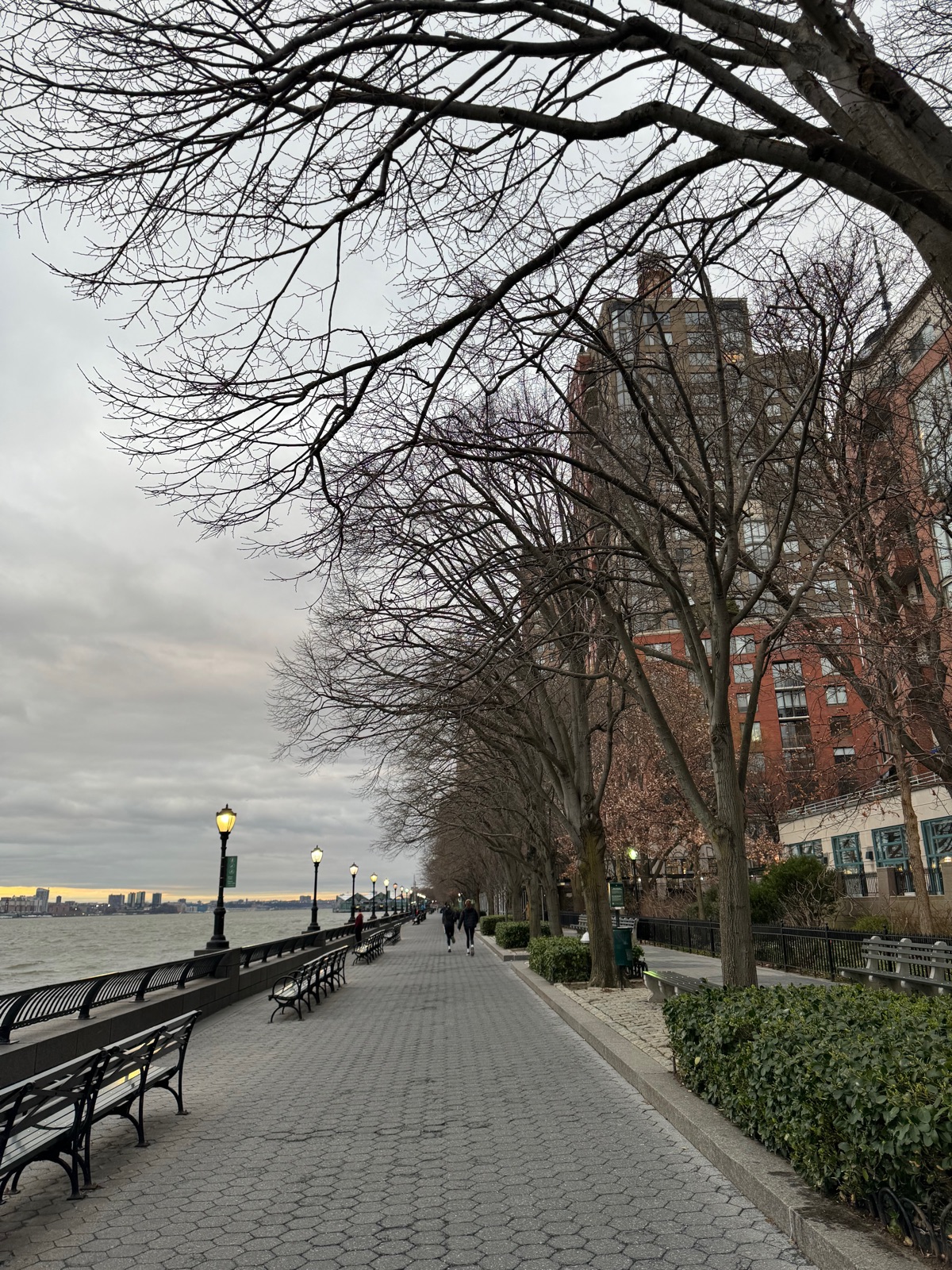 A wide paved esplanade lined with benches and bare trees running along the Hudson at dusk, two distant figures walking away