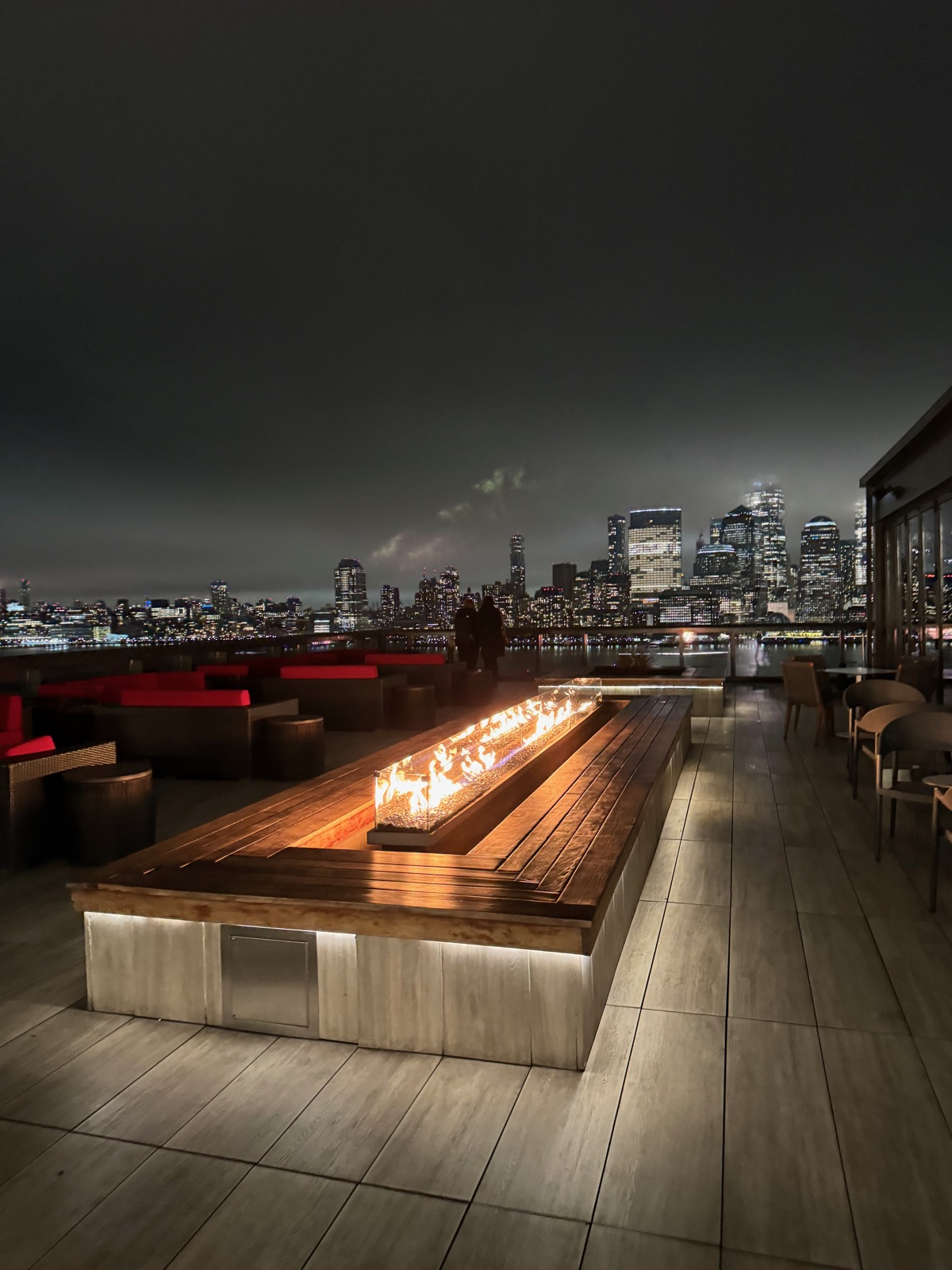 A long gas fire trough burning down the middle of a timber terrace at night, with lounge seating around it and the lit Lower Manhattan skyline across the black water beyond