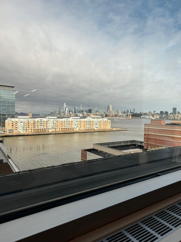 The Manhattan skyline framed through a window from the New Jersey side, a low waterfront apartment complex in the middle ground