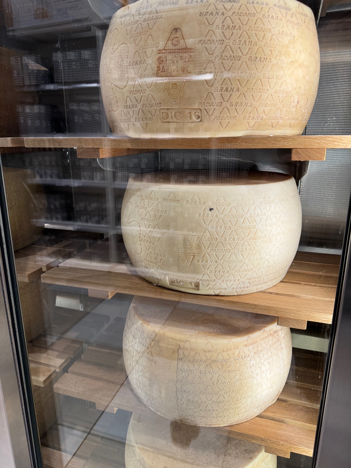 Three whole wheels of Grana Padano stacked on oak shelves behind glass, the rinds stamped repeatedly with the name and the DOP mark