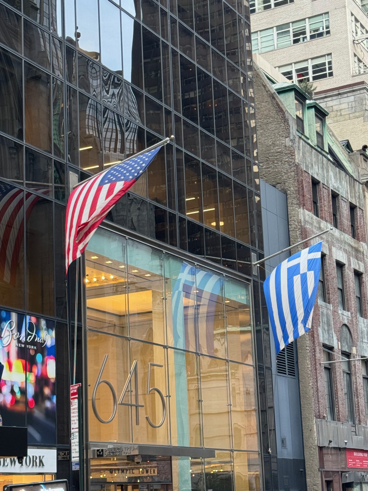 A Greek flag and a United States flag hanging side by side at the same height from poles on the glass front of a Fifth Avenue building, with the number 645 on the glass below
