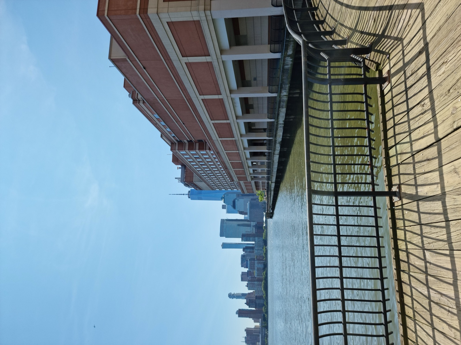 A railing along the Hudson with the Manhattan skyline low on the horizon across the water under a clear blue sky
