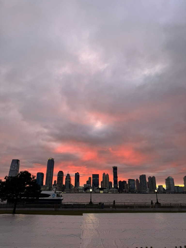 A dramatic pink and coral sunset over the Hudson River, the skyline in silhouette across the water, a ferry passing