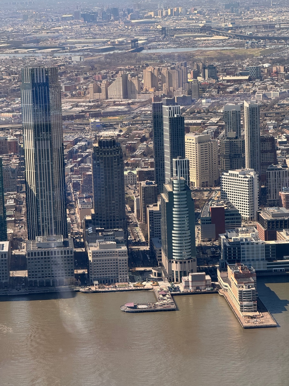 The Jersey City waterfront and its towers seen from high above across the brown Hudson, with piers and low industrial buildings behind