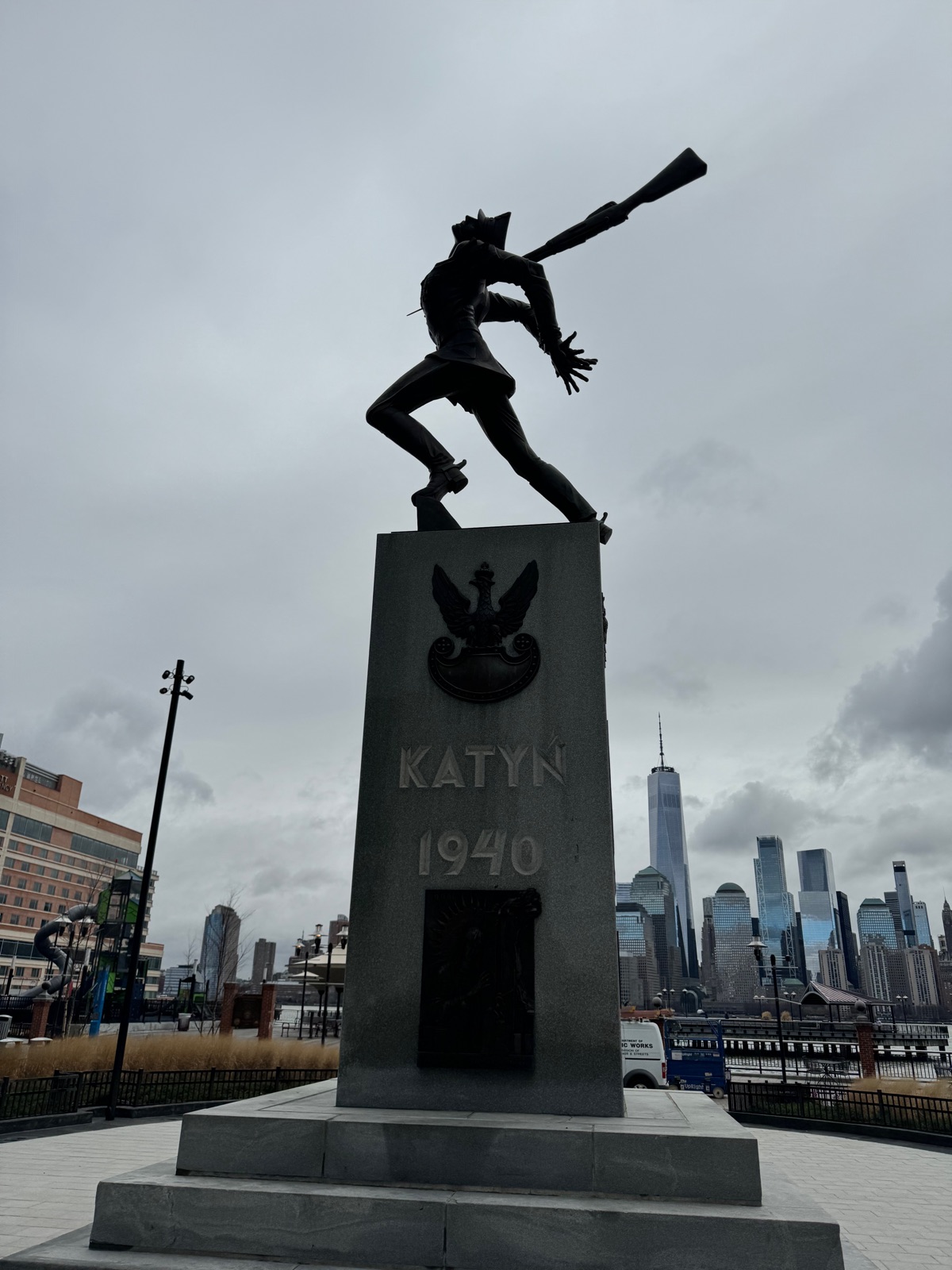 A bronze statue of a Polish soldier falling forward with a bayonet through his back, on a stone pedestal reading KATYŃ 1940, with the Lower Manhattan skyline across the water behind it
