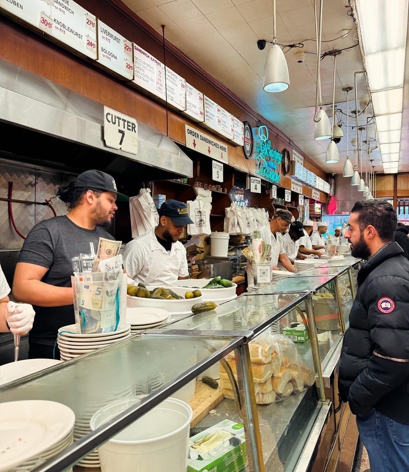 The counter at Katz's Delicatessen, cutters in white shirts and caps slicing meat under numbered station signs, a customer ordering