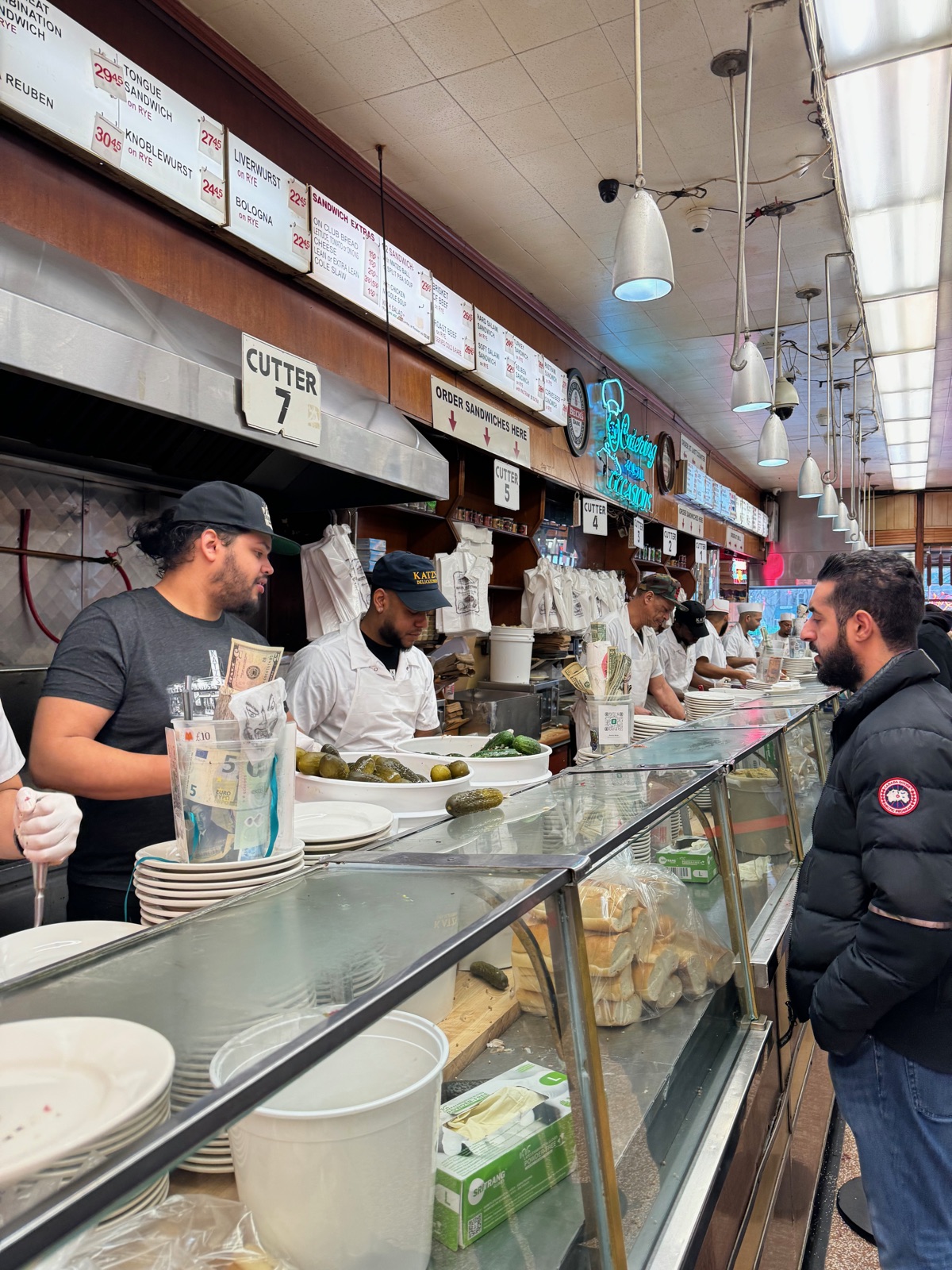 The carving counter at Katz's Delicatessen with a sign reading CUTTER 7 above it, staff in caps working behind the glass case, stacked plates, bowls of pickles and a cup of dollar bills