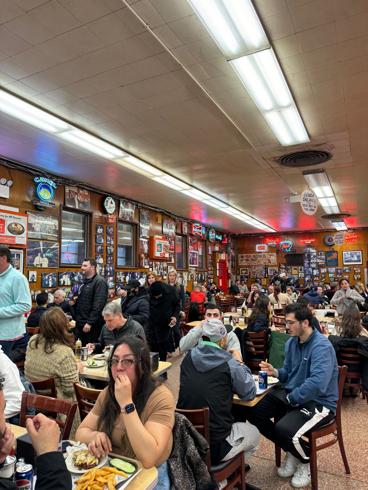 The crowded dining room at Katz's Delicatessen, rows of tables full of people under strip lights and neon beer signs, photographs covering the walls and a hanging sign reading SEND A SALAMI TO YOUR BOY IN THE ARMY