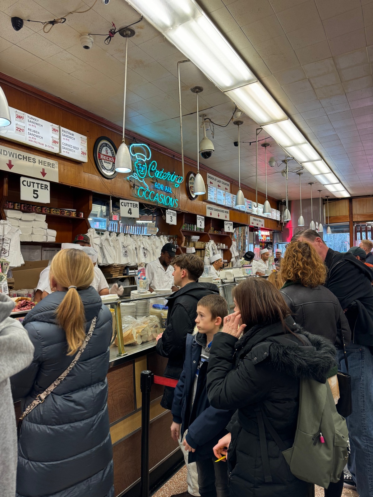 People queueing at the counter inside Katz's Delicatessen beneath hanging lamps and menu boards, staff in white behind the glass case