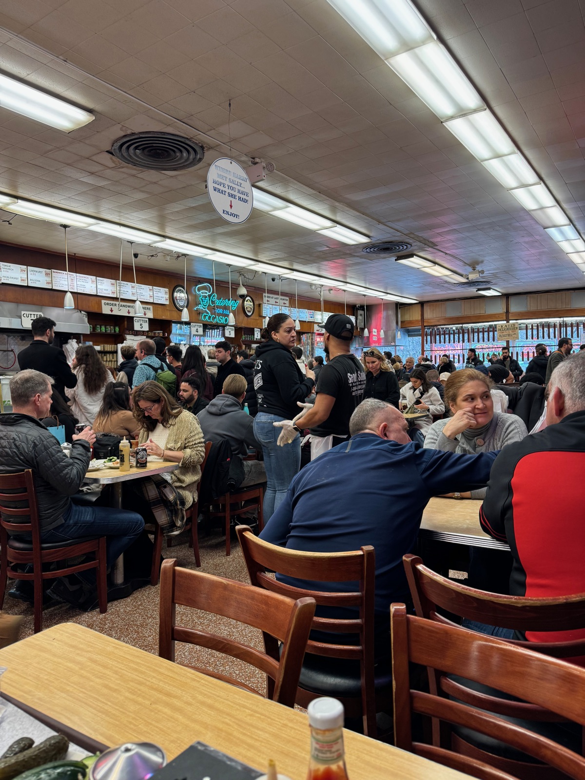 A wide view across the packed tables at Katz's Delicatessen, a round sign hanging from the ceiling reading WHERE HARRY MET SALLY, HOPE YOU HAVE WHAT SHE HAD, menu boards and neon on the far wall, and pickles and a bottle on the table in front