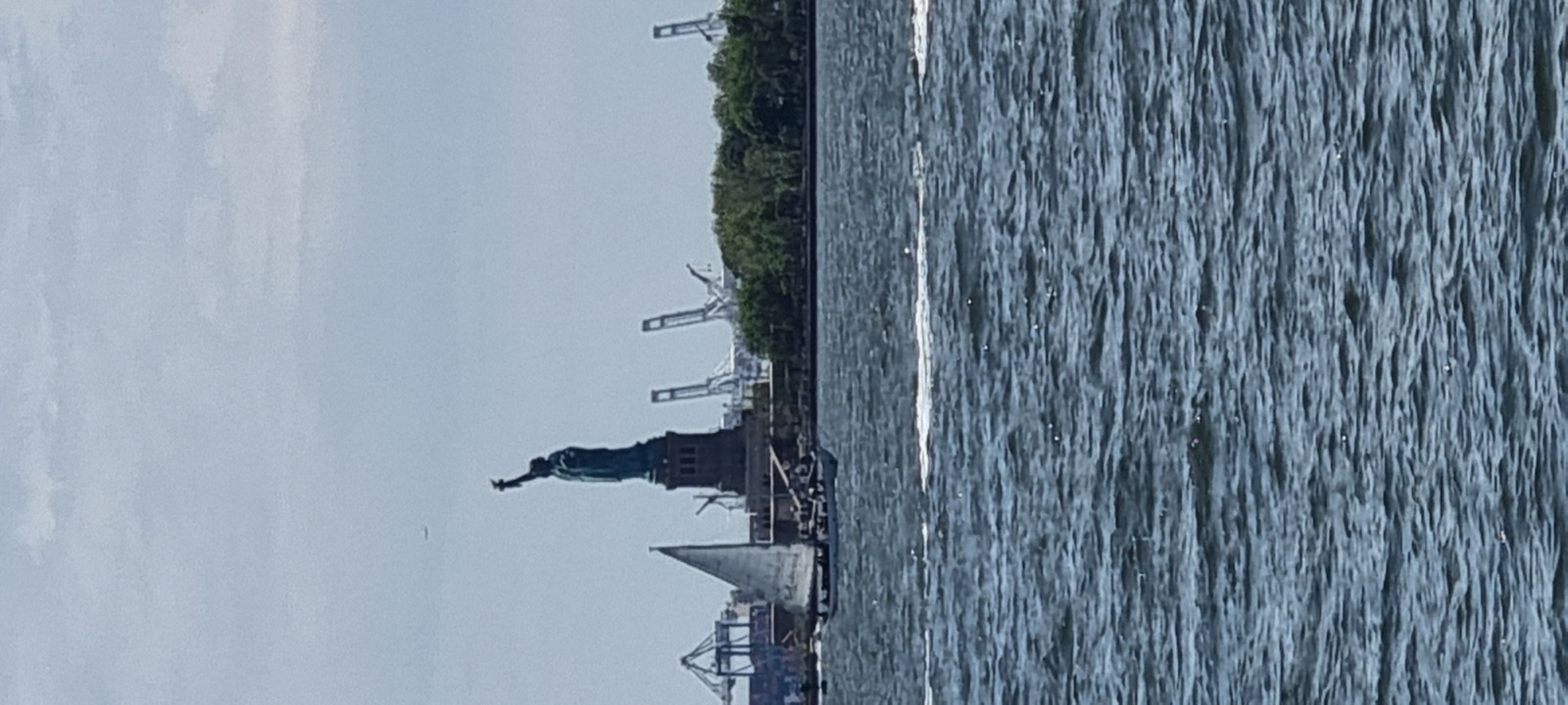 The Statue of Liberty small and distant across the harbor under a pale sky, with cranes and low industrial buildings on the far shore