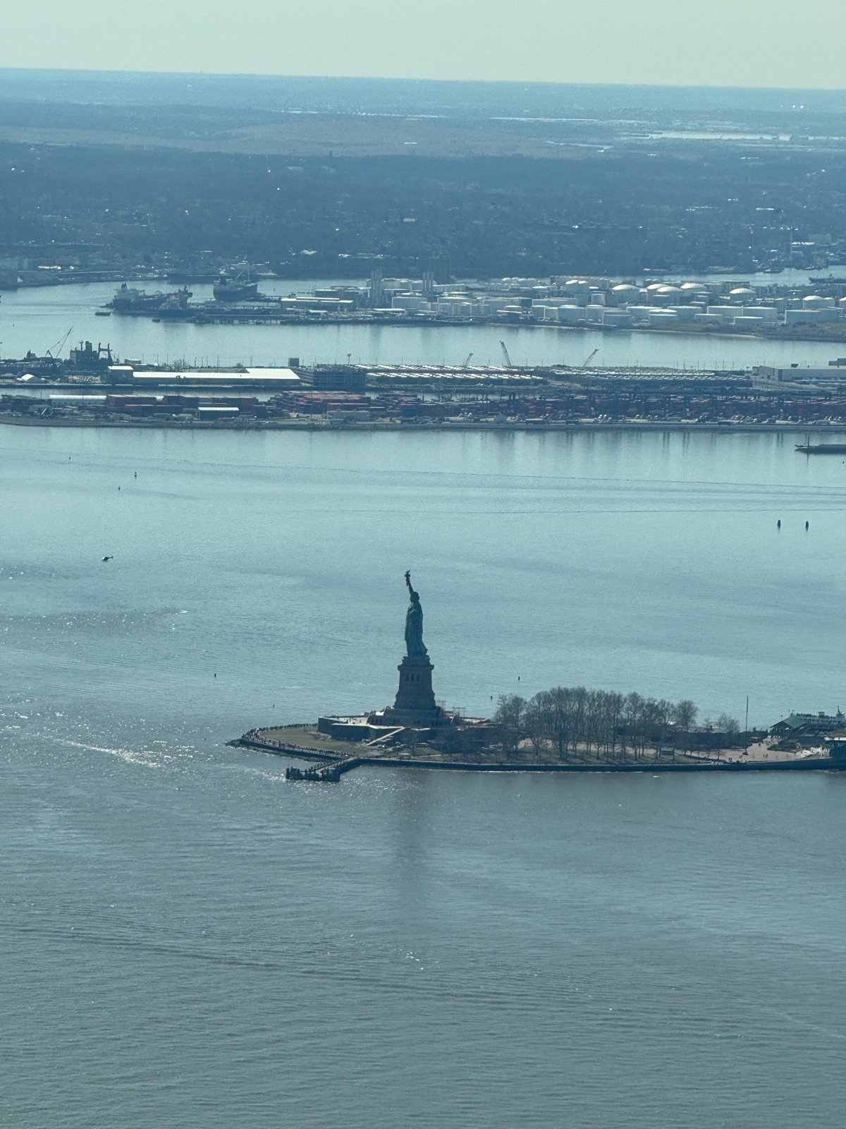 The Statue of Liberty on its island in the middle of a wide gray-blue harbor, seen from high above