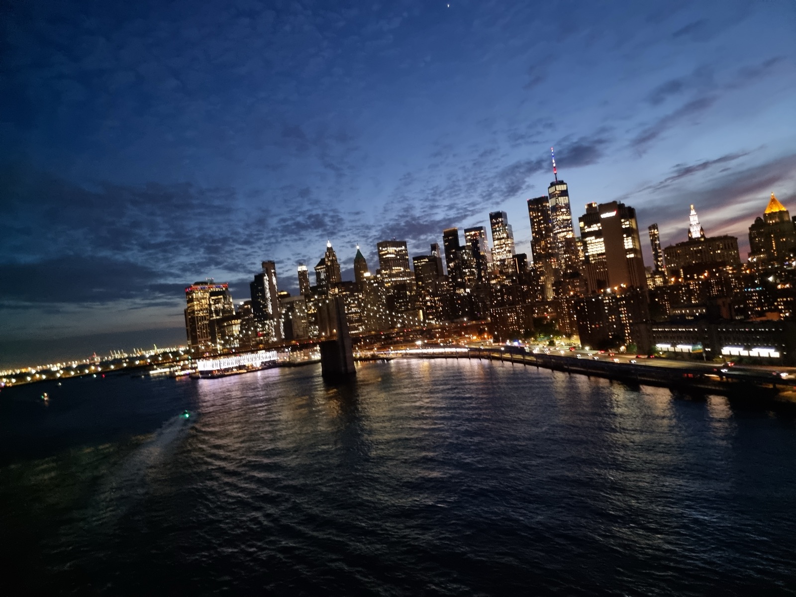 The Lower Manhattan skyline lit at blue hour seen across the East River, the water dark and the sky still holding color