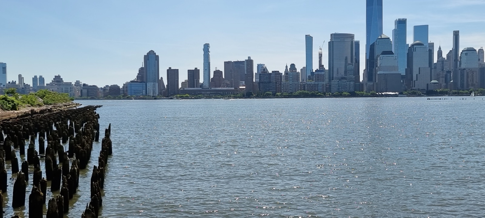 The full Lower Manhattan skyline seen across a calm Hudson from the Jersey shore, with a row of piling stumps in the foreground