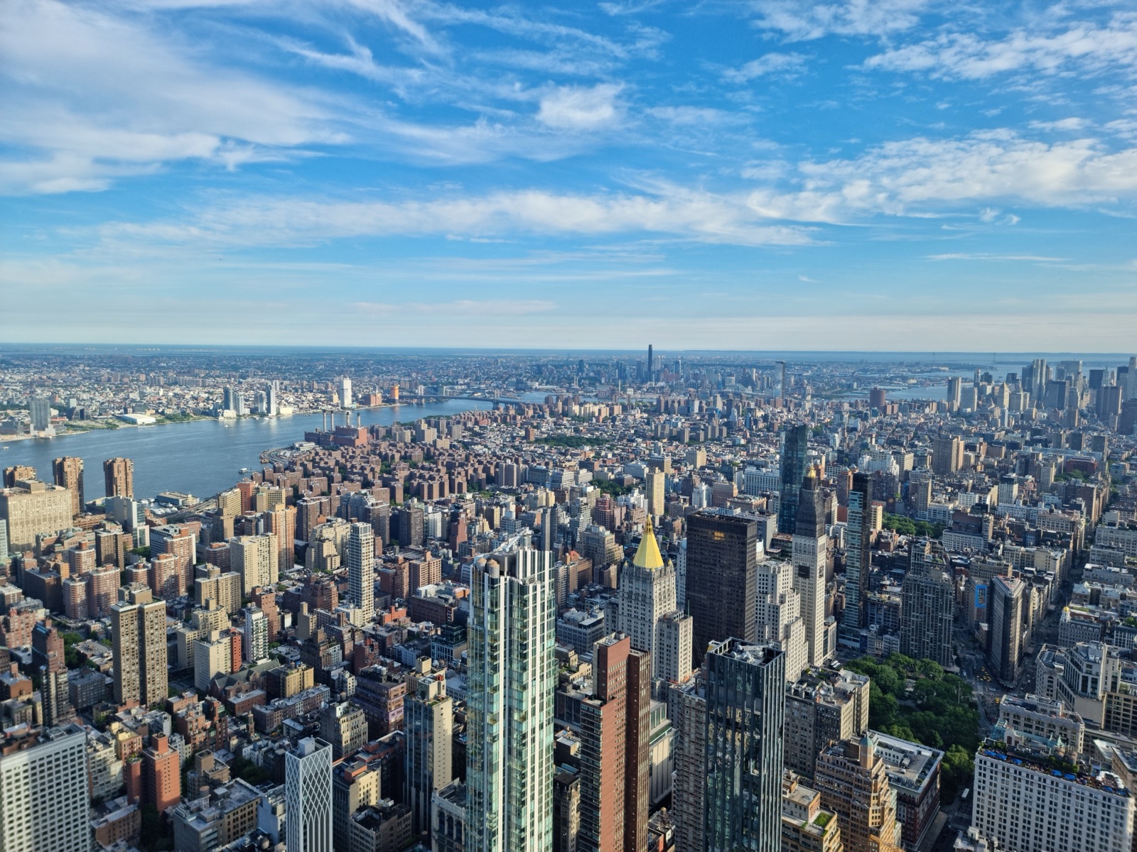 A wide view north over the Manhattan grid from the Empire State Building, dense towers running to the rivers on both sides under a blue sky with high cloud