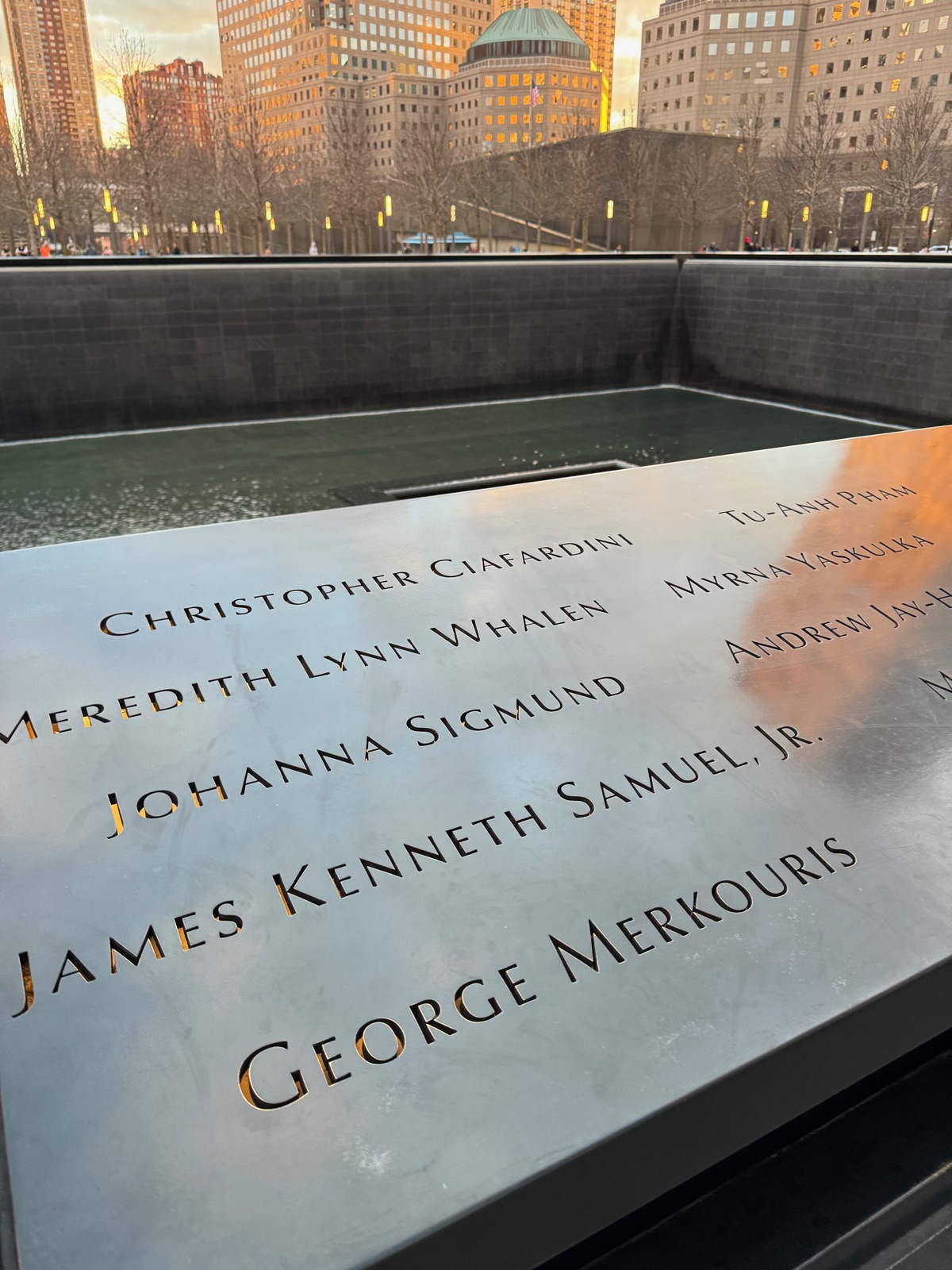 Names cut through the bronze parapet of a September 11 memorial pool in the late afternoon sun, reading Christopher Ciafardini, Meredith Lynn Whalen, Johanna Sigmund, James Kenneth Samuel Jr and George Merkouris