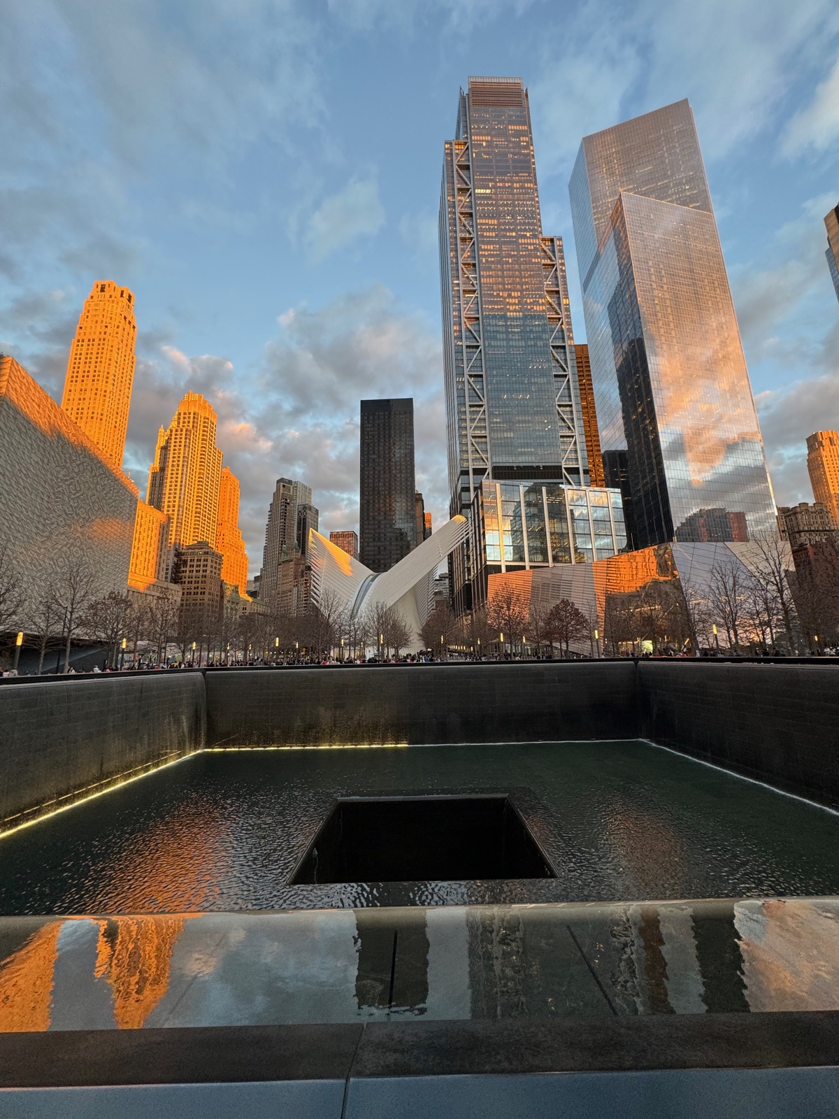 A memorial pool with the surrounding towers lit orange by the setting sun and reflected in the wet granite