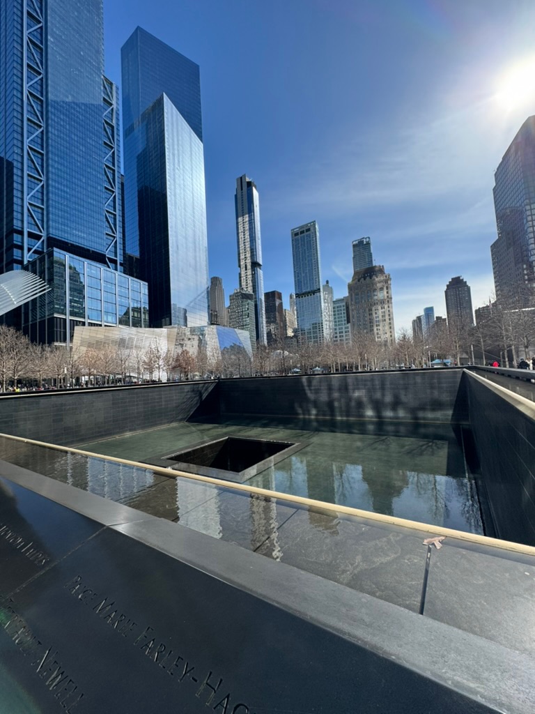 The September 11 Memorial reflecting pool by day, water running down black granite, names engraved on the bronze parapet