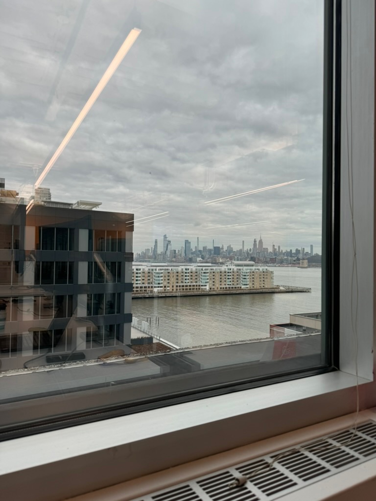 The Midtown Manhattan skyline in the distance across the Hudson, seen through an office window with aircraft contrails overhead
