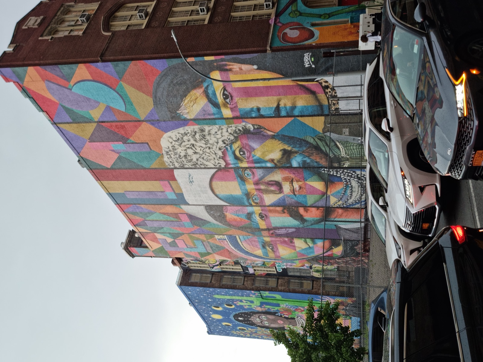 A large brightly colored mural of overlapping human faces painted across the whole side wall of a brick building, cars parked in front of it