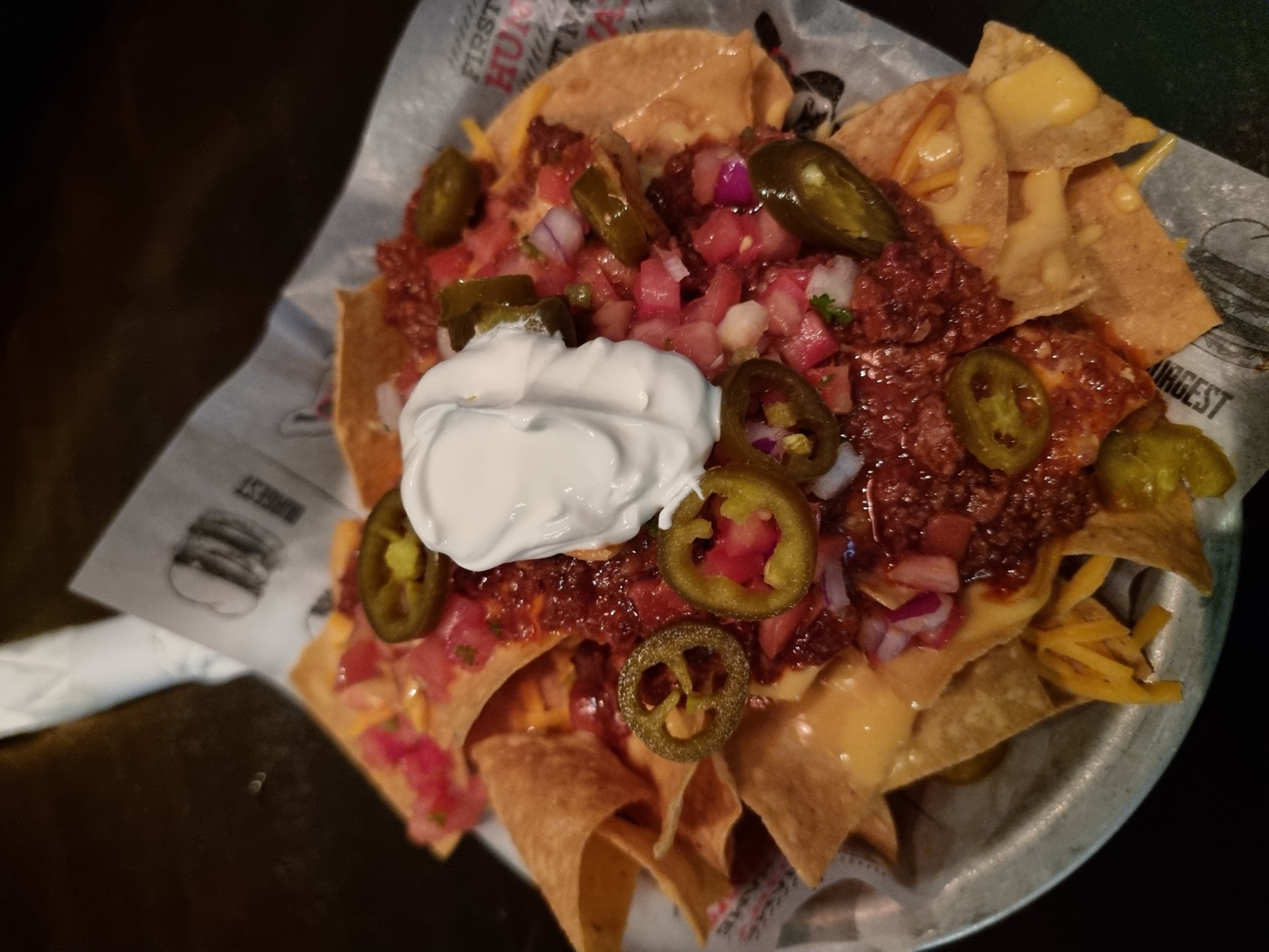 A dish of nachos loaded with chili, melted cheese, sliced jalapeños, diced tomato and a scoop of sour cream