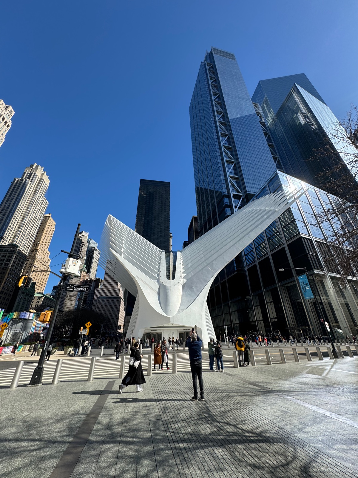 The white winged exterior of the Oculus rising between dark glass towers, people crossing the sunlit plaza in front of it