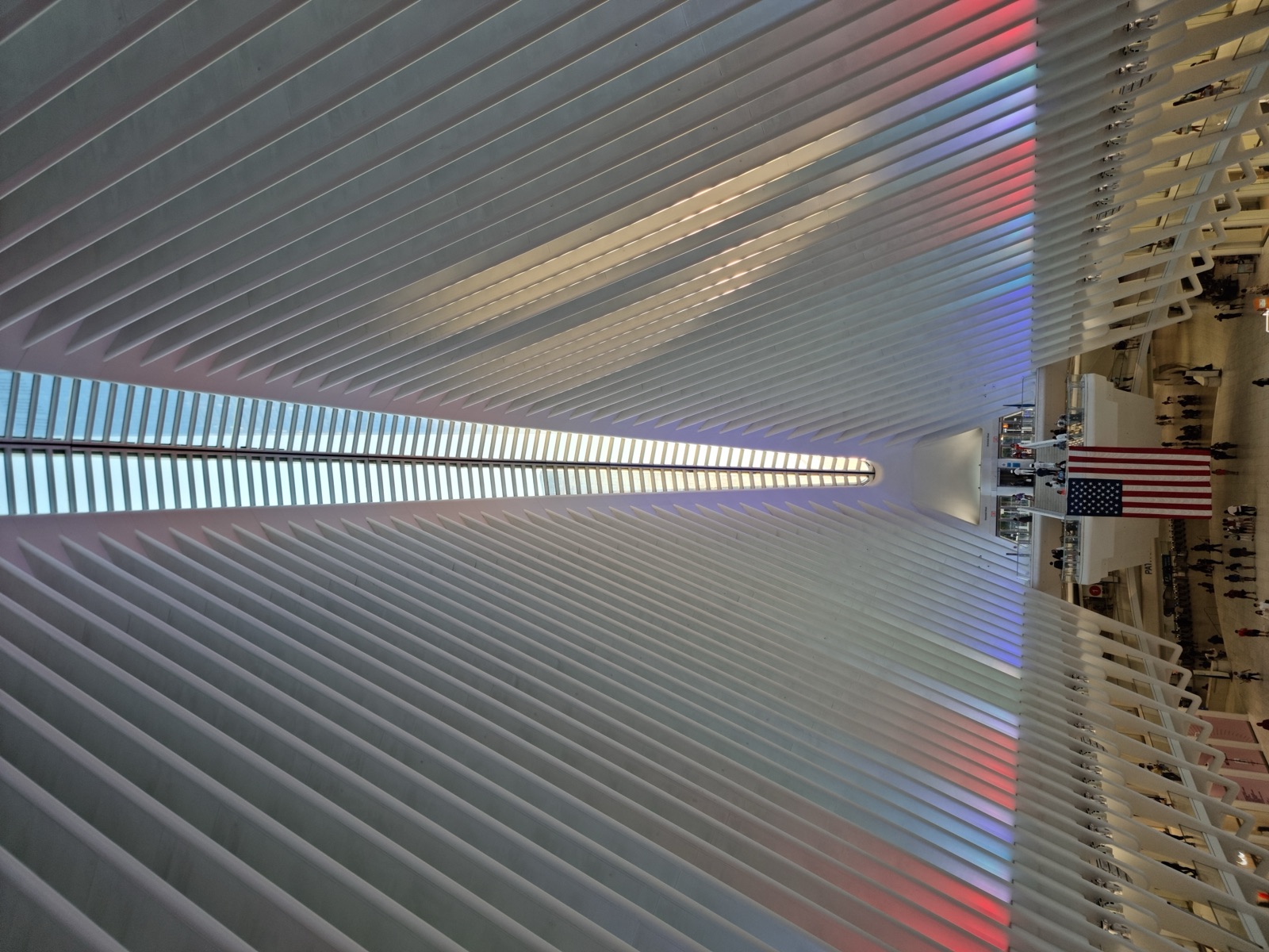The interior of the Oculus at night with its white ribs lit in red and blue along their length, converging on the bright skylight, a large United States flag hanging over the concourse below