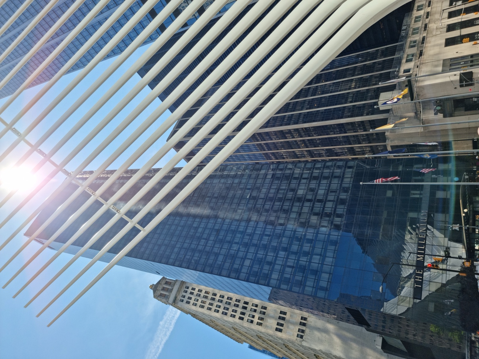 The white ribbed wing of the Oculus seen from below at a steep angle with the sun flaring between the ribs and a dark glass tower behind