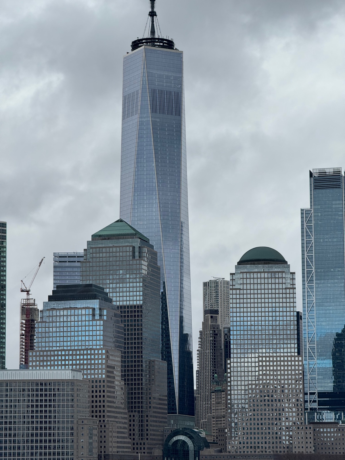One World Trade Center rising above the Lower Manhattan skyline under heavy gray cloud, seen from across the Hudson