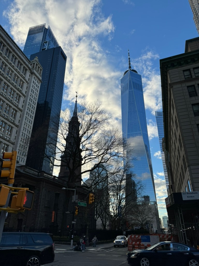 One World Trade Center rising behind the stone steeple of Saint Paul's Chapel, seen from Broadway under a blue sky