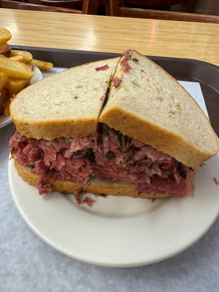 A hand-cut pastrami sandwich piled high on rye, served on a white plate with a side of fries