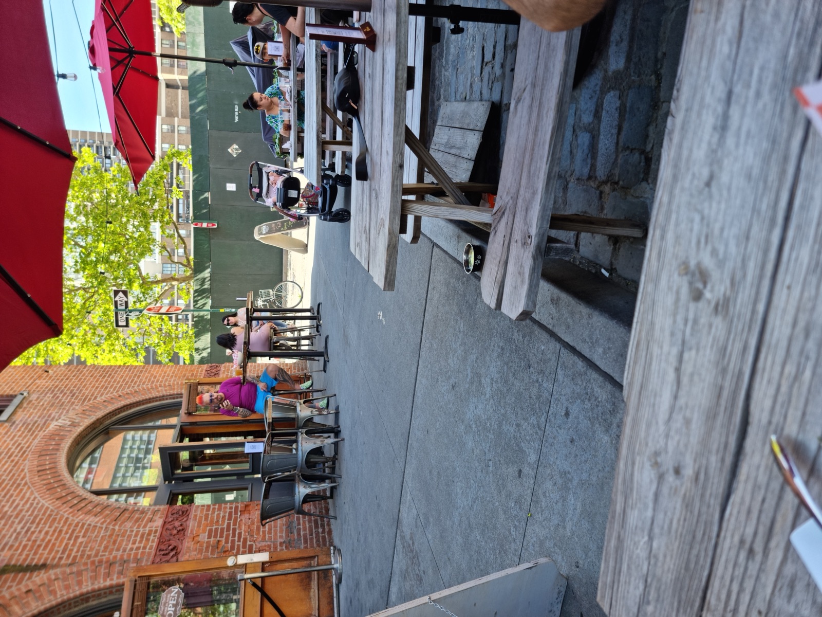 Weathered wooden picnic tables under red umbrellas outside a brick warehouse building, with people at the far tables