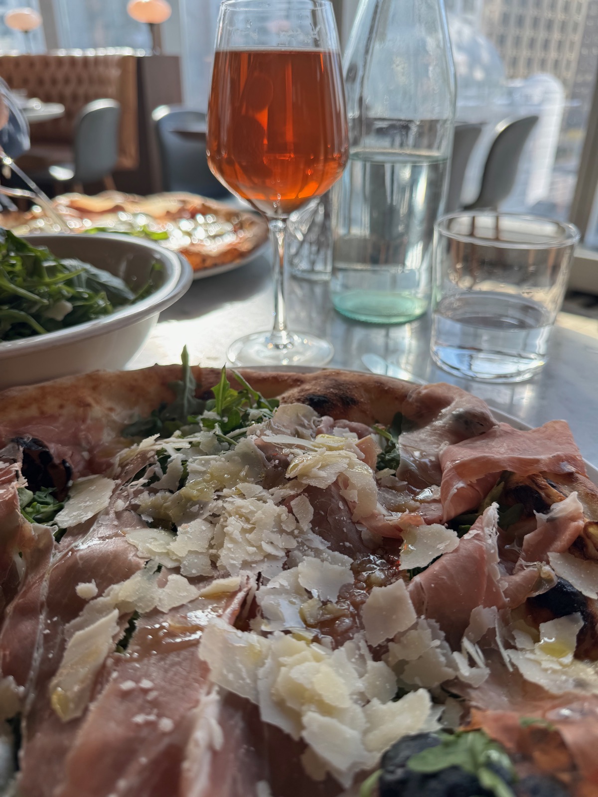 A pizza topped with prosciutto, arugula and shaved cheese on a table, with a glass of amber drink and a bowl of salad behind it
