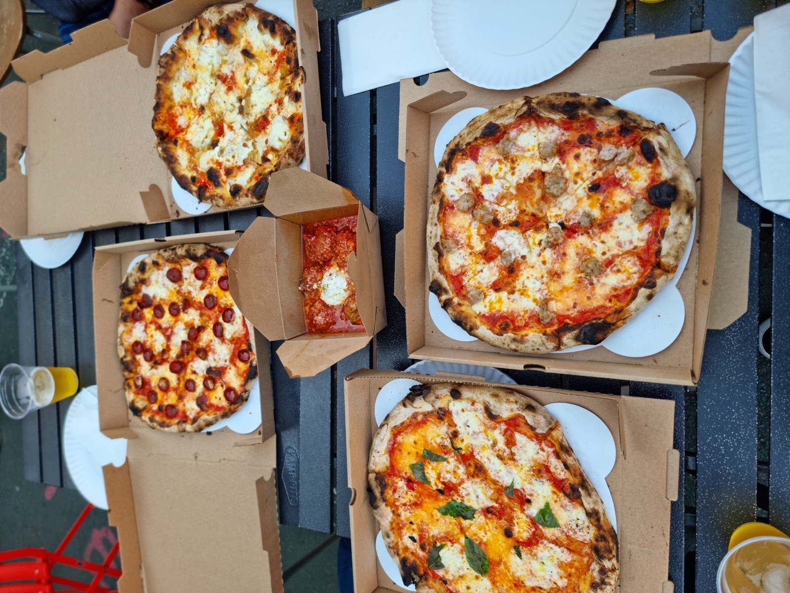 Four Neapolitan pizzas open in their cardboard boxes on a dark picnic table with paper plates and a glass of beer