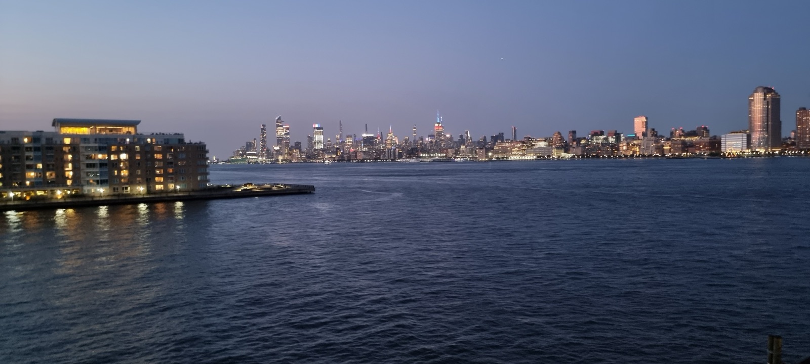 The Manhattan skyline across the Hudson at dusk with the water pale and the sky deepening to blue above the towers