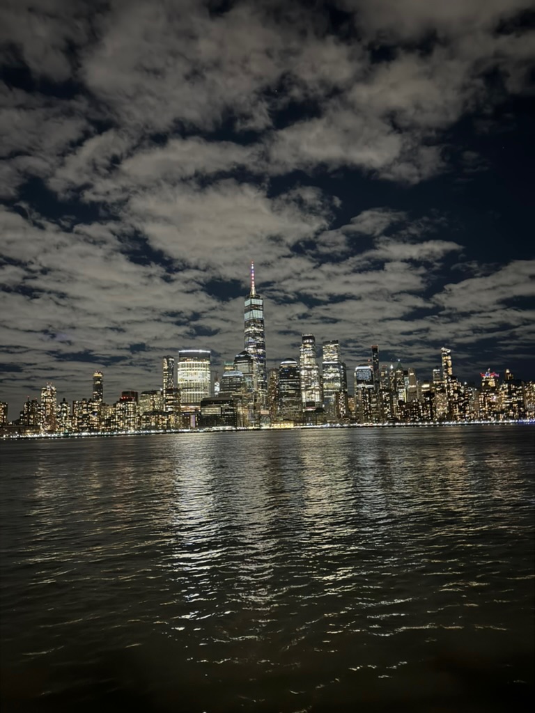 Lower Manhattan skyline at night under dramatic moonlit clouds, One World Trade lit red at its tip, reflections on the water